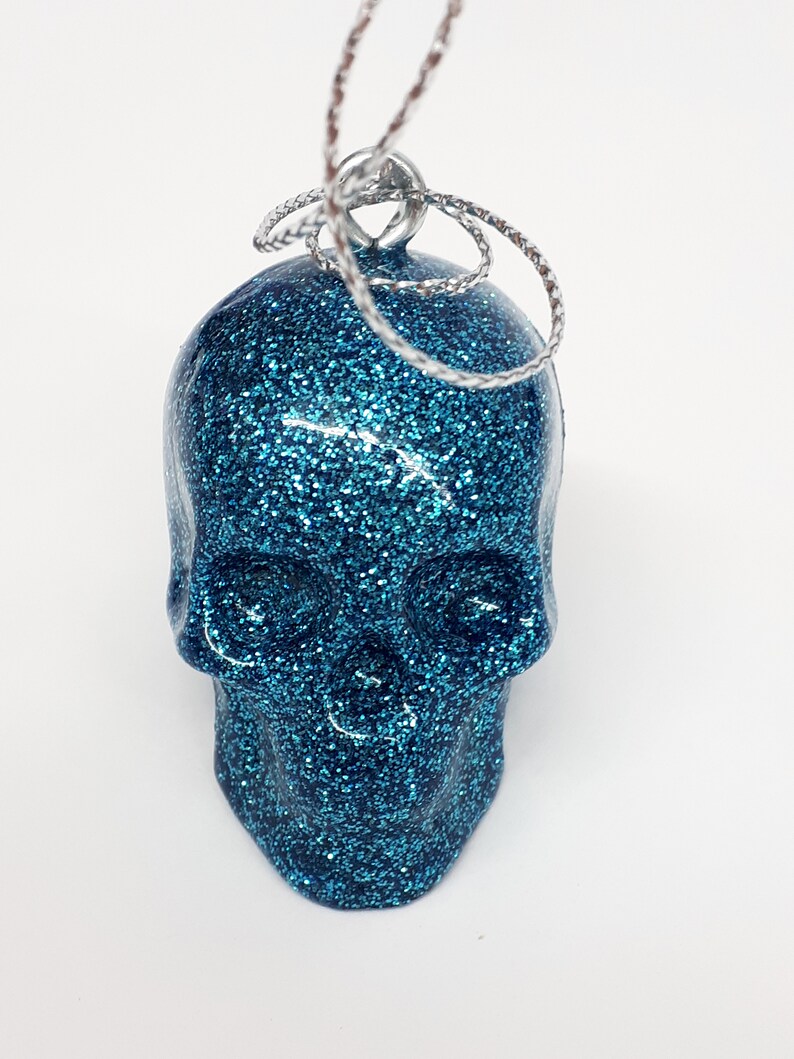Puede incluir: Un adorno de calavera azul con purpurina y una cuerda plateada para colgar.