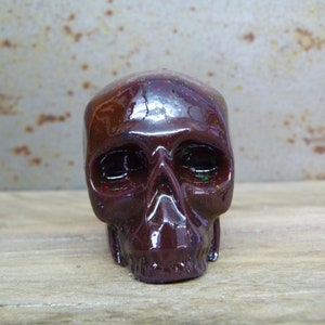 May include: A small, dark red skull with a glossy finish and iridescent glitter embedded in the resin. The skull has a detailed face with eye sockets, a nose, and a mouth.