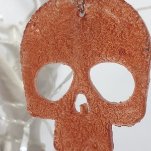May include: A copper-colored skull-shaped ornament with a detailed, textured surface. The ornament is suspended by a silver chain. The skull has large eye sockets and a nose cavity, and is likely a Halloween or gothic-themed decoration.