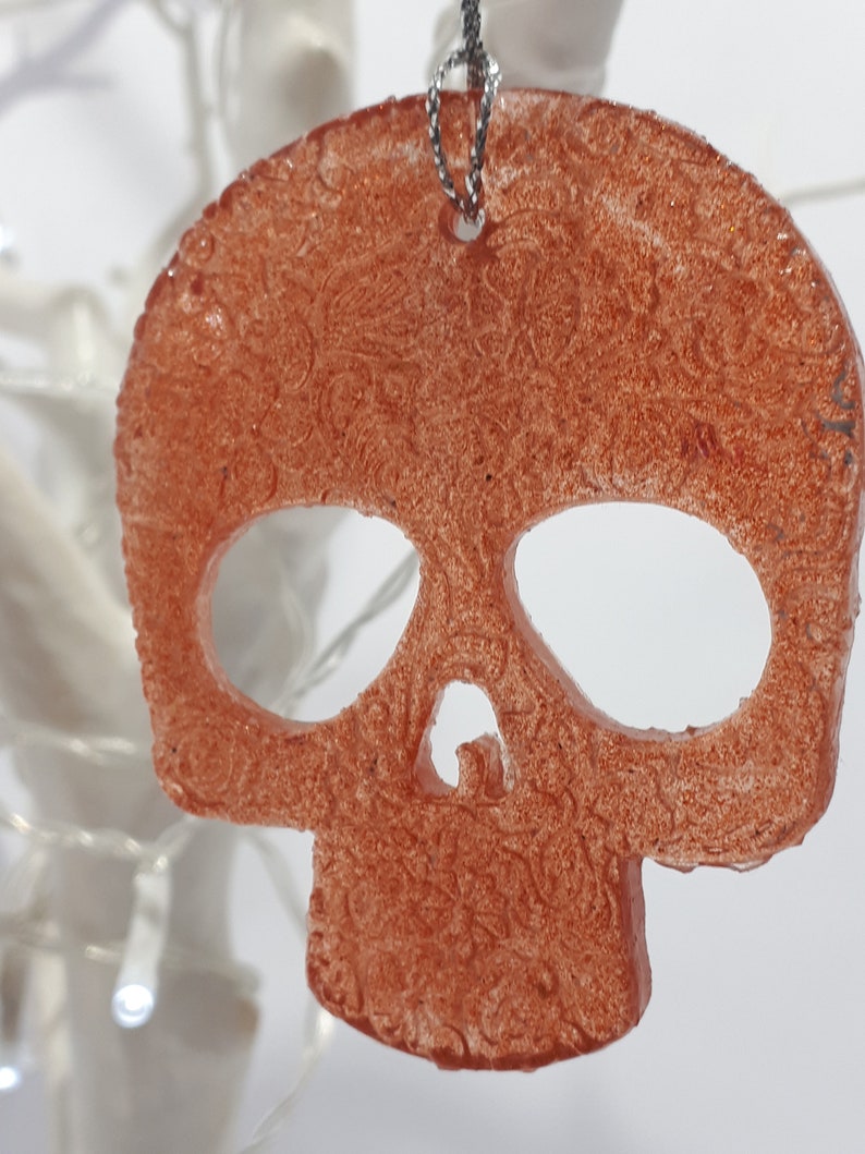 May include: A copper-colored skull-shaped ornament with a detailed, textured surface. The ornament is suspended by a silver chain. The skull has large eye sockets and a nose cavity, and is likely a Halloween or gothic-themed decoration.