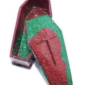 Pu&ograve; includere: Una scatola a forma di bara rossa e verde con glitter e una croce sul coperchio. La scatola &egrave; aperta e il coperchio &egrave; appoggiato sopra la scatola.