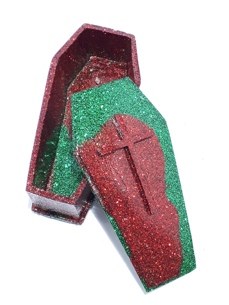 Pu&ograve; includere: Una scatola a forma di bara rossa e verde con glitter e una croce sul coperchio. La scatola &egrave; aperta e il coperchio &egrave; appoggiato sopra la scatola.