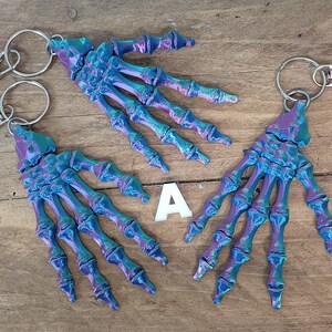 May include: Three iridescent blue and purple skeleton hand keyrings with silver key rings. The keychains are on a wooden surface.