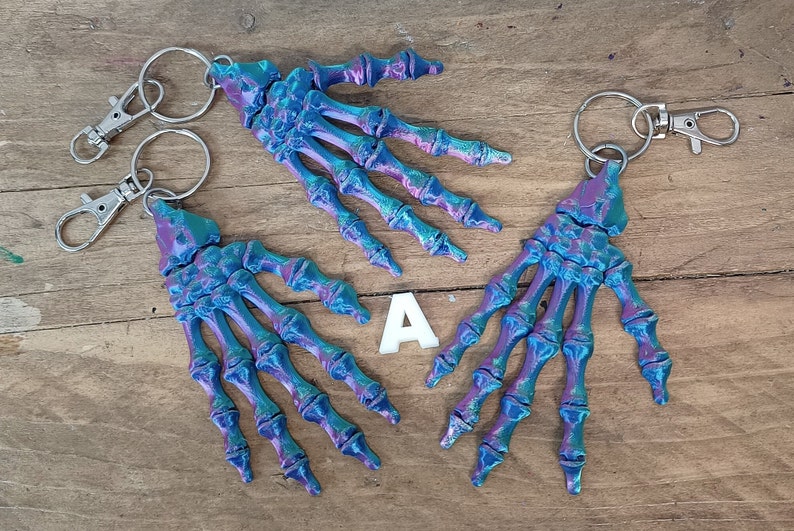 May include: Three iridescent blue and purple skeleton hand keyrings with silver key rings. The keychains are on a wooden surface.