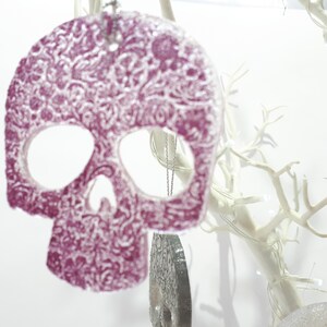 May include: A decorative, purple skull ornament with a detailed floral pattern. The skull has cut-out eye and nose sockets and hangs from a silver chain. The ornament is displayed against a white background with white branches.