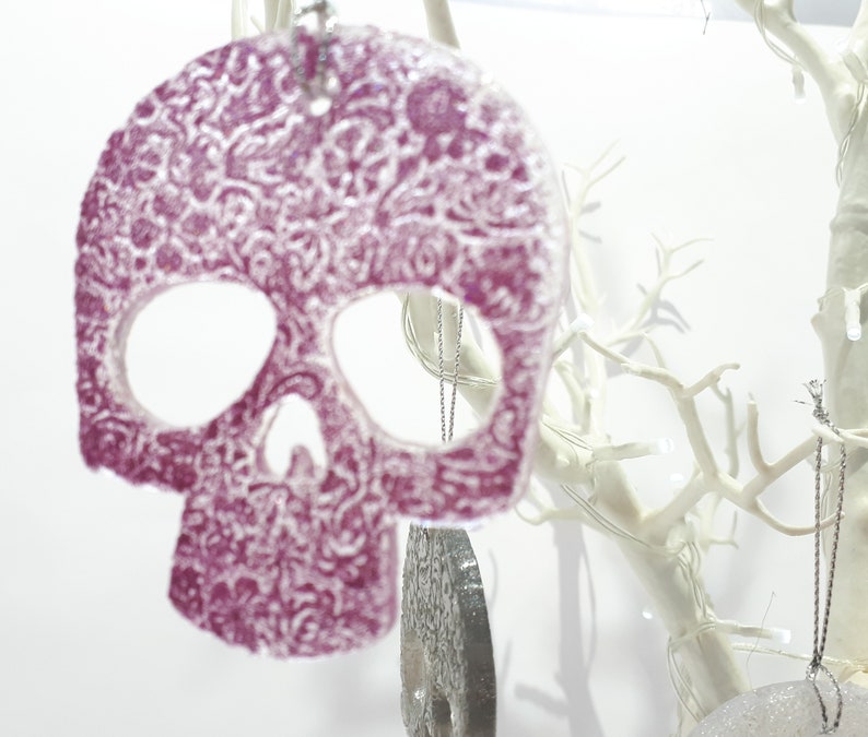 May include: A decorative, purple skull ornament with a detailed floral pattern. The skull has cut-out eye and nose sockets and hangs from a silver chain. The ornament is displayed against a white background with white branches.