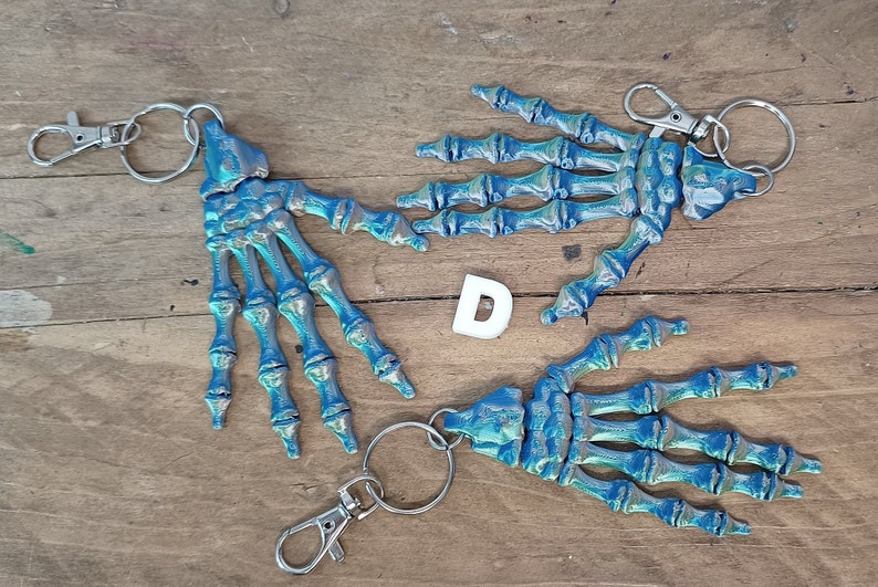 May include: Three skeleton hand keyrings with silver key rings. The hands are painted in a blue and silver iridescent colour.