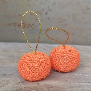 K&ouml;nnte beinhalten: Zwei orangefarbene H&auml;kelornamente mit goldenen Drahtschlaufen. Die Ornamente sind rund und haben eine zarte, spitzenartige Textur.