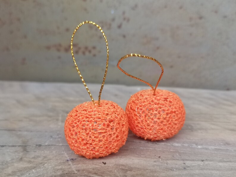 K&ouml;nnte beinhalten: Zwei orangefarbene H&auml;kelornamente mit goldenen Drahtschlaufen. Die Ornamente sind rund und haben eine zarte, spitzenartige Textur.