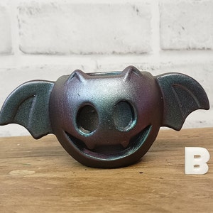 May include: A black and iridescent ceramic bat-shaped candle holder with a smiling face and wings. The holder is sitting on a wooden surface.