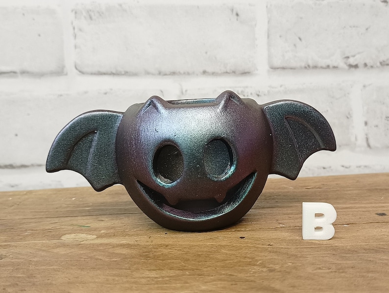 May include: A black and iridescent ceramic bat-shaped candle holder with a smiling face and wings. The holder is sitting on a wooden surface.