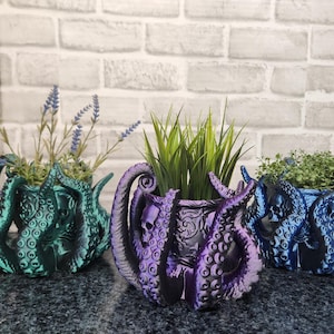 May include: Three octopus-shaped planters in teal, purple, and blue, each containing greenery. The planters have tentacle designs and skull details. The teal planter holds lavender, the purple planter holds tall grass, and the blue planter holds small green plants.