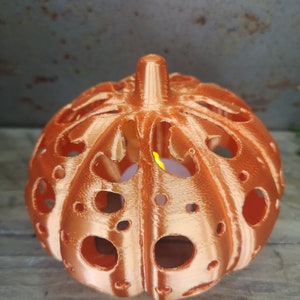 May include: A copper-colored pumpkin-shaped candle holder with a decorative, intricate design and cut-out holes. The candle holder is lit with a small candle inside.