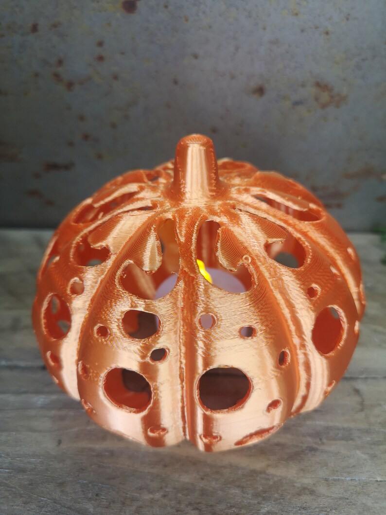 May include: A copper-colored pumpkin-shaped candle holder with a decorative, intricate design and cut-out holes. The candle holder is lit with a small candle inside.