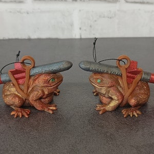 May include: Two brown frog ornaments with red and black missile ornaments on their backs. The frogs are sitting on a gray surface.