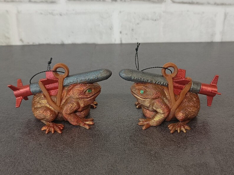 May include: Two brown frog ornaments with red and black missile ornaments on their backs. The frogs are sitting on a gray surface.