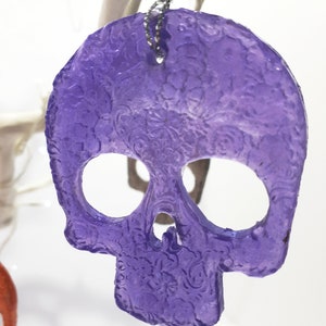 May include: A translucent purple skull-shaped ornament with floral detailing. The skull has large eye sockets and a nose cavity. A silver chain is attached for hanging. The ornament is likely for Halloween or gothic decor.