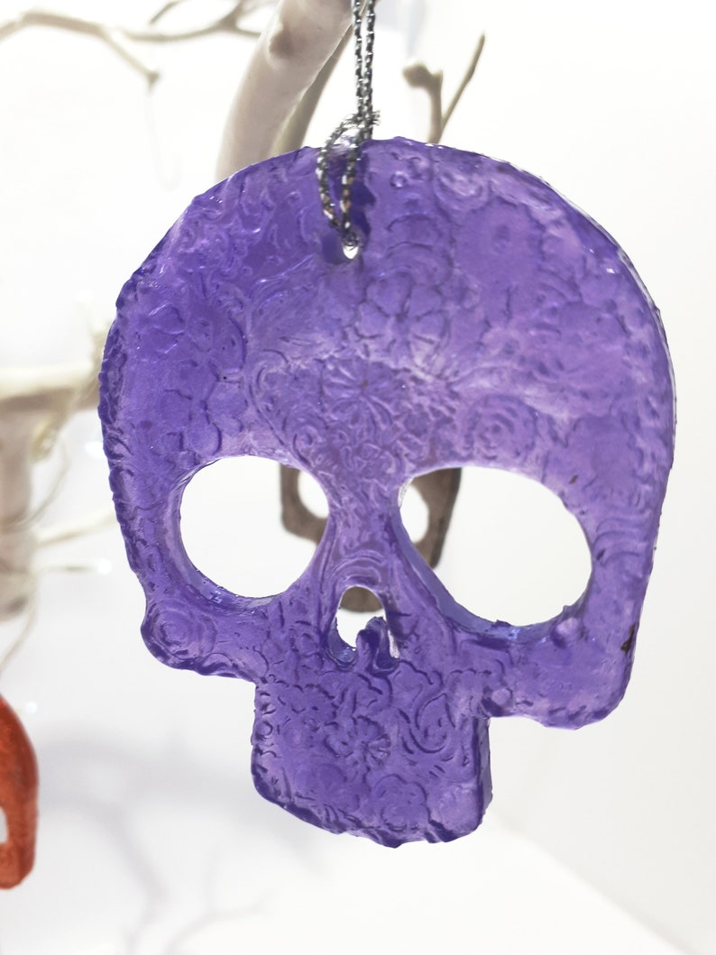 May include: A translucent purple skull-shaped ornament with floral detailing. The skull has large eye sockets and a nose cavity. A silver chain is attached for hanging. The ornament is likely for Halloween or gothic decor.