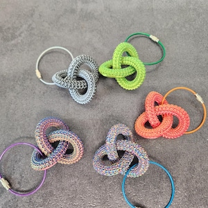 Puede incluir: Cinco llaveros coloridos, anudados en forma de anillo, con anillos metálicos. Los llaveros están hechos de un material texturizado similar al plástico. Los colores son verde, rojo, gris, azul y morado.