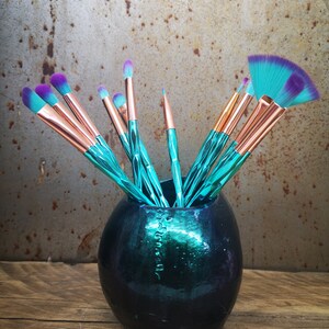 K&ouml;nnte beinhalten: Eine tealfarbene und schwarze Keramikvase mit einem Satz Make-up-Pinsel mit tealfarbenen und violetten Griffen. Die Pinsel sind in der Vase angeordnet, wobei die Griffe herausragen.