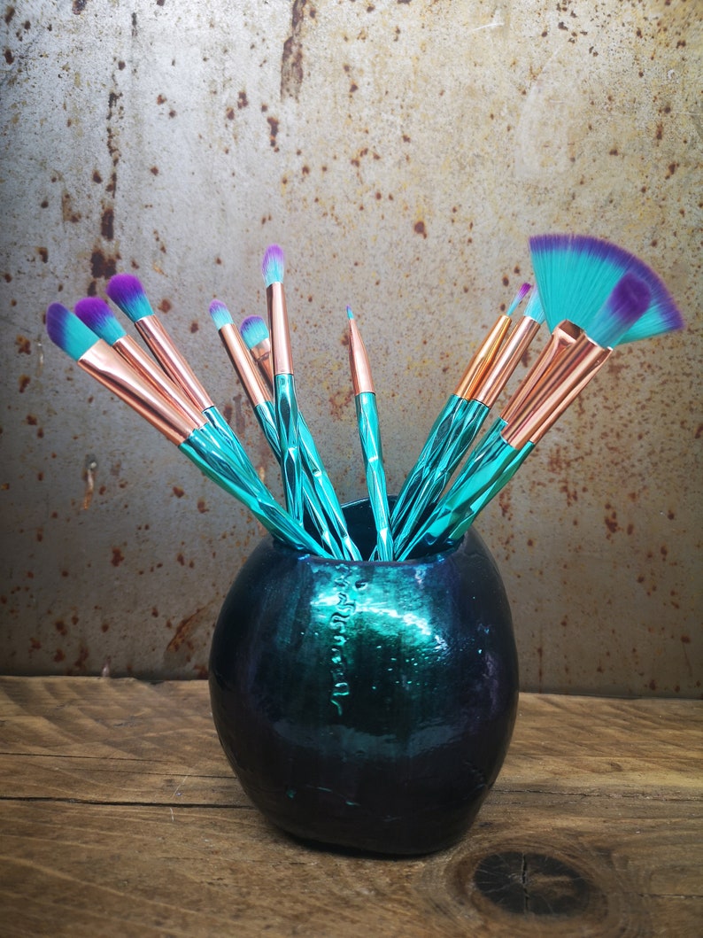 K&ouml;nnte beinhalten: Eine tealfarbene und schwarze Keramikvase mit einem Satz Make-up-Pinsel mit tealfarbenen und violetten Griffen. Die Pinsel sind in der Vase angeordnet, wobei die Griffe herausragen.