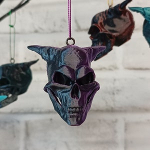 May include: Several hanging skull ornaments with a dark, iridescent finish. The skulls have horns and are suspended by purple cords. The background is a white brick wall. These skulls are a unique decorative item.