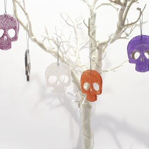 May include: A collection of skull-shaped ornaments in various colors, including purple, orange, and white, hanging from a white decorative tree. The ornaments have a textured surface and are suspended by silver strings. The background is plain white.
