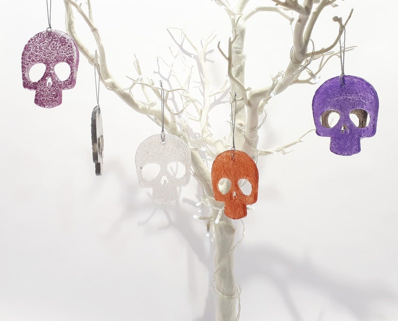 May include: A collection of skull-shaped ornaments in various colors, including purple, orange, and white, hanging from a white decorative tree. The ornaments have a textured surface and are suspended by silver strings. The background is plain white.