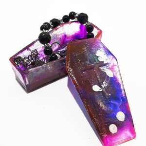 Pu&ograve; includere: Una scatola per gioielli a forma di bara di colore viola e rosa con un braccialetto di perline nere e una catena d'argento all'interno. La scatola &egrave; aperta e il coperchio &egrave; sdraiato di lato.