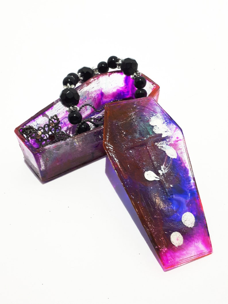 Pu&ograve; includere: Una scatola per gioielli a forma di bara di colore viola e rosa con un braccialetto di perline nere e una catena d'argento all'interno. La scatola &egrave; aperta e il coperchio &egrave; sdraiato di lato.