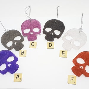 May include: Six skull-shaped ornaments in various colors: purple, gray, glittery pink, black, white, and red. Each ornament has a silver string for hanging. Scrabble tiles with letters and numbers are placed near each skull.
