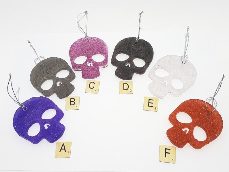 May include: Six skull-shaped ornaments in various colors: purple, gray, glittery pink, black, white, and red. Each ornament has a silver string for hanging. Scrabble tiles with letters and numbers are placed near each skull.