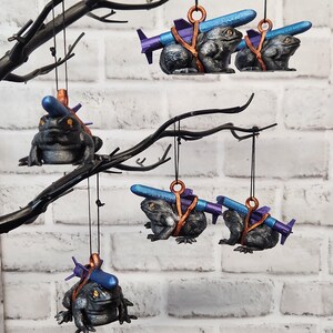 May include: Five black frog ornaments hanging from a black branch. Each frog is holding a blue and purple missile. The frogs are wearing brown straps. The ornaments are hanging from a white brick wall.