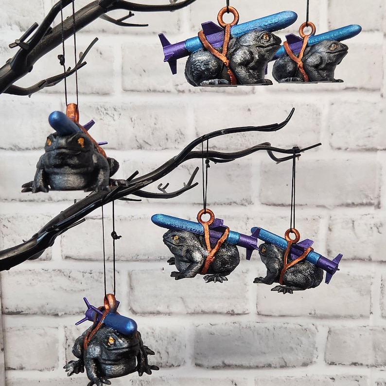 May include: Five black frog ornaments hanging from a black branch. Each frog is holding a blue and purple missile. The frogs are wearing brown straps. The ornaments are hanging from a white brick wall.