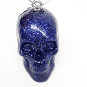 Puede incluir: Un colgante de calavera azul con purpurina en una cadena plateada.