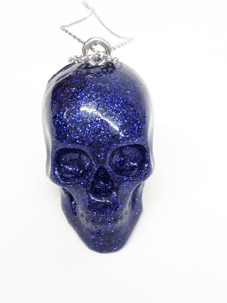 Puede incluir: Un colgante de calavera azul con purpurina en una cadena plateada.