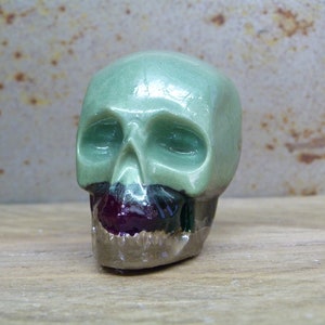 May include: A small ceramic skull sculpture with a green glaze on the top half and a black and burgundy glaze on the bottom half. The skull has a detailed face with eye sockets and a mouth.