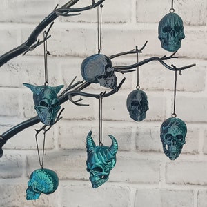 May include: A set of six teal green skull ornaments hanging from a black branch. The skulls are all different designs, including a skull with horns, a skull with a spiral design, and a skull with a detailed face.