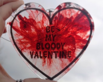 May include: A clear heart-shaped ornament with a red and white marbled design. The ornament has a black outline and the text "Be My Bloody Valentine" written in black.