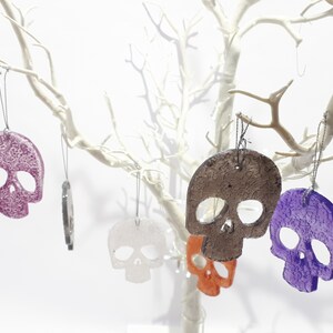 May include: Several skull-shaped ornaments in various colors, including purple, orange, and white, hang from the branches of a white decorative tree. Each ornament has a textured surface and is suspended by a thin string, creating a spooky yet festive display.