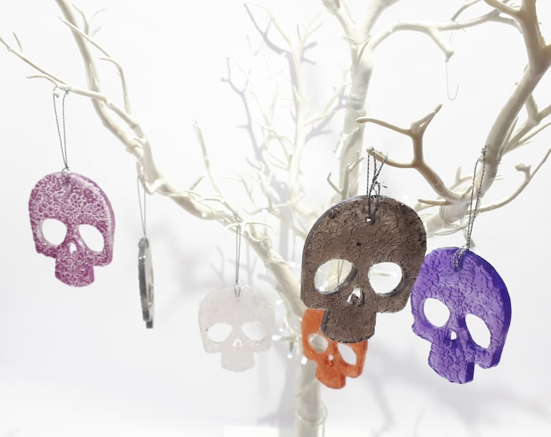May include: Several skull-shaped ornaments in various colors, including purple, orange, and white, hang from the branches of a white decorative tree. Each ornament has a textured surface and is suspended by a thin string, creating a spooky yet festive display.
