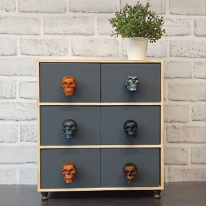 May include: A wooden chest of drawers with six drawers, each with a skull-shaped knob. The drawers are painted a dark grey colour. The chest of drawers is sitting on a white brick wall.