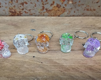 May include: Six clear resin skull keychains with different colored pressed flowers inside. The keychains are on a wooden surface.