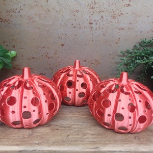 May include: Three red, hollow, pumpkin-shaped candle holders with a dotted pattern. The candle holders are made of a smooth, shiny material and are sitting on a wooden surface.