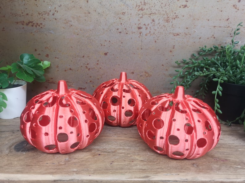 May include: Three red, hollow, pumpkin-shaped candle holders with a dotted pattern. The candle holders are made of a smooth, shiny material and are sitting on a wooden surface.