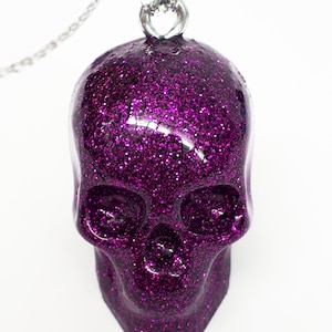 Puede incluir: Un colgante de calavera de purpurina morada en una cadena plateada.