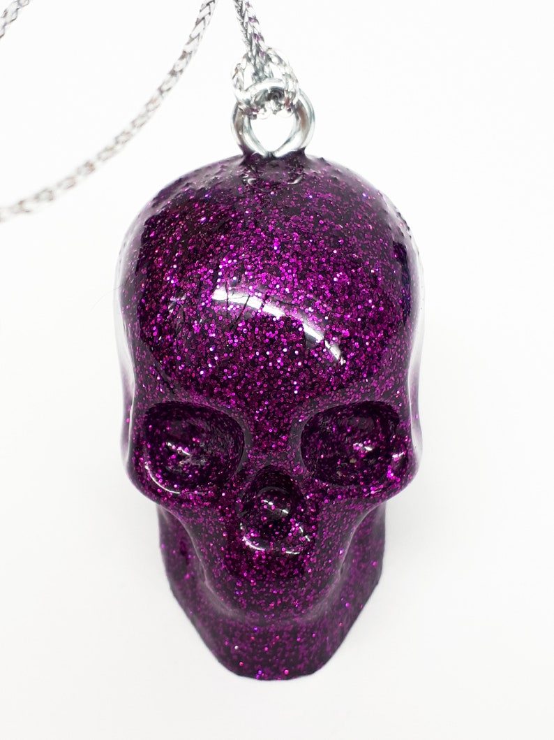 Puede incluir: Un colgante de calavera de purpurina morada en una cadena plateada.