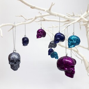 Puede incluir: Una colecci&oacute;n de adornos de calavera con purpurina en varios colores, incluyendo plata, morado, azul y turquesa, colgando de una rama blanca.
