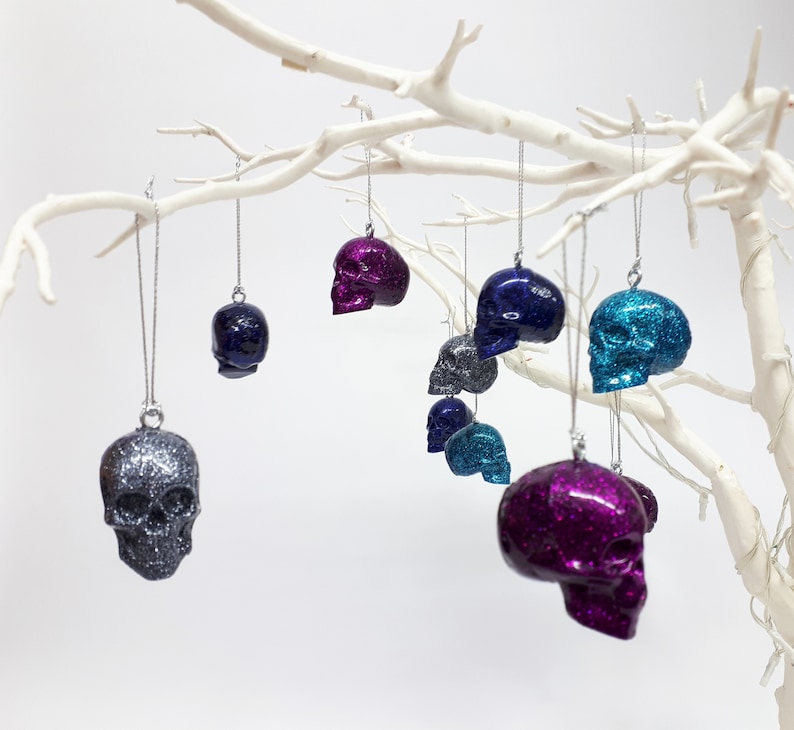 Puede incluir: Una colecci&oacute;n de adornos de calavera con purpurina en varios colores, incluyendo plata, morado, azul y turquesa, colgando de una rama blanca.