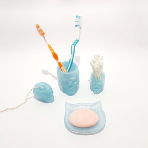 May include: A blue ceramic skull-shaped toothbrush holder with two toothbrushes and a blue ceramic skull-shaped cotton swab holder with cotton swabs. There is also a blue ceramic soap dish with a pink bar of soap.